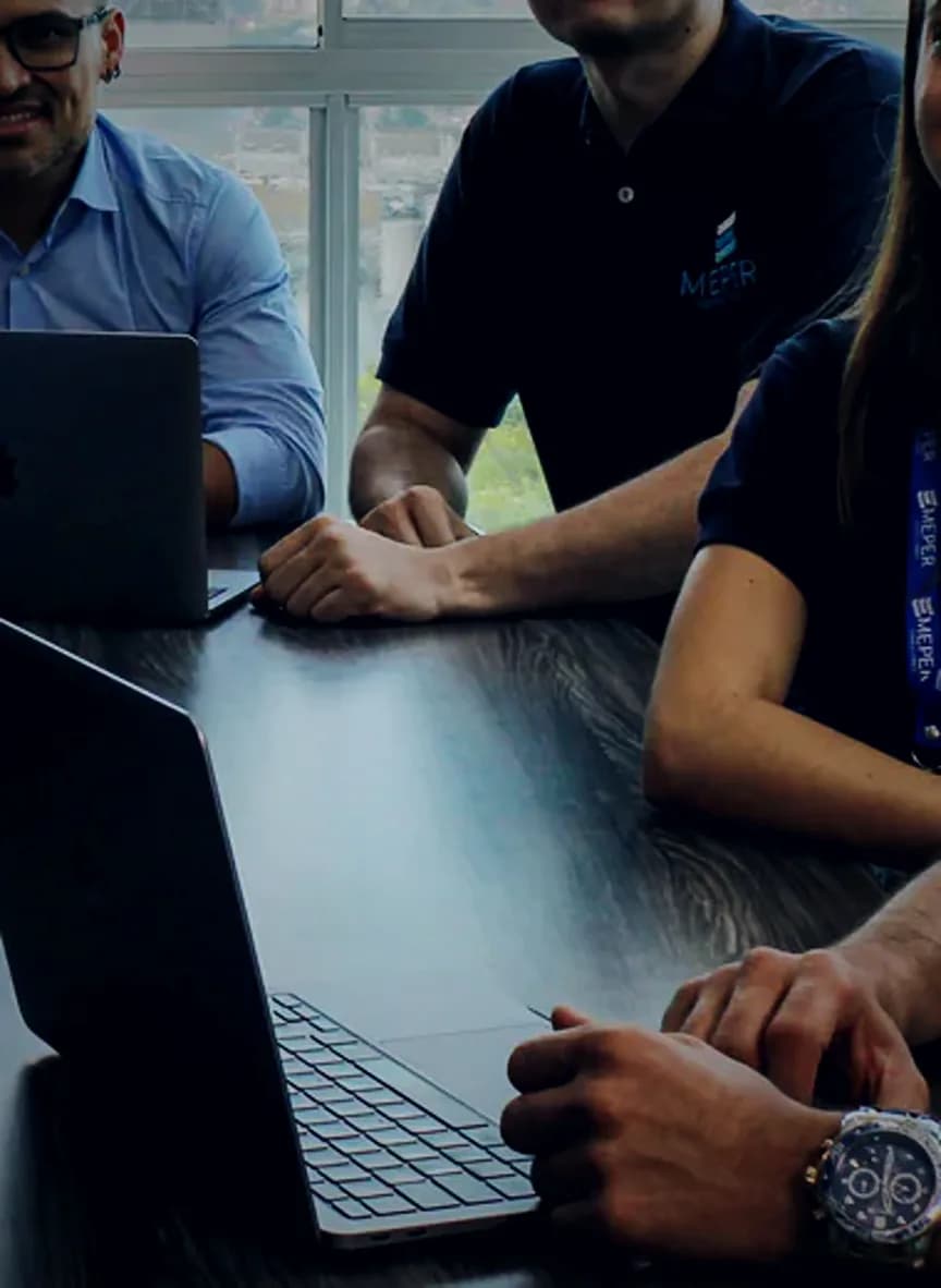 Equipo de MEPER Consultores trabajando con portátiles abiertos en sala de juntas, con la cordillera de Medellín al fondo