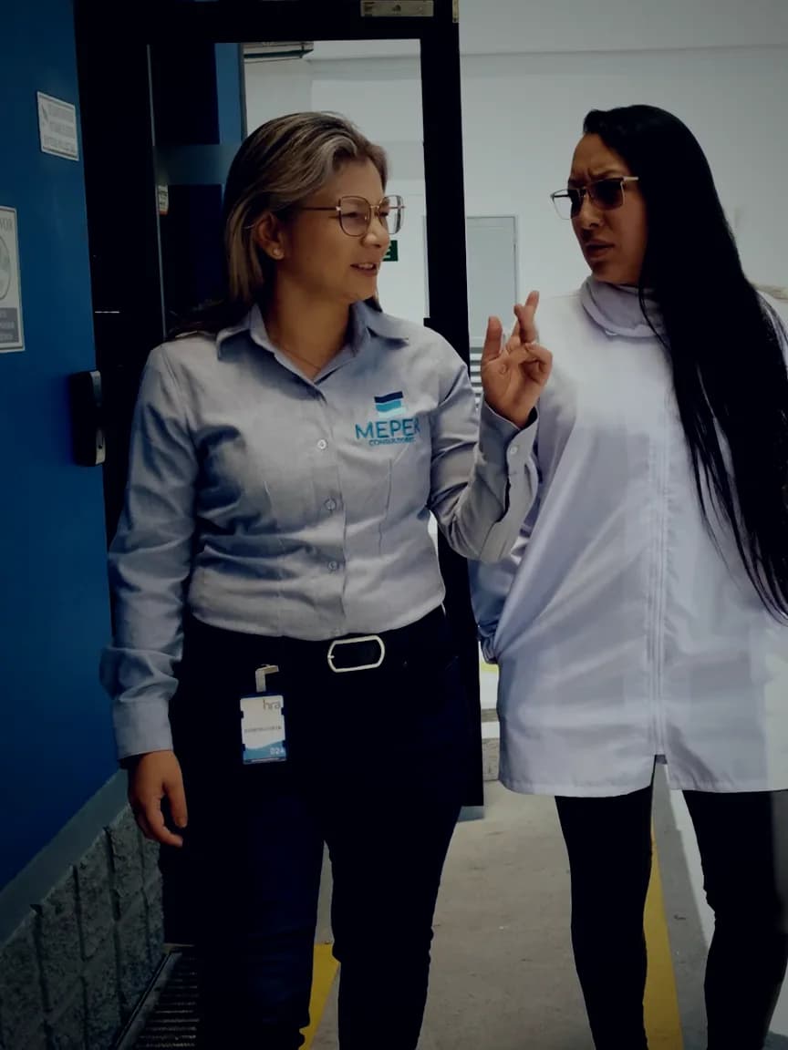Dos consultoras de MEPER caminando y conversando en una bodega industrial, con un montacargas detrás